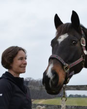 Dr. Amy Johnson standing outdoors smiling at a dark brown horse wearing a halter with a cloudy sky and wooden fence in the background.
