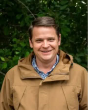 Assoc. Prof. Steven Zedler smiling outdoors in a tan jacket, standing in front of lush greenery.