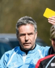Dr. Bruce Bladon wearing a light blue sports shirt, holding up a yellow card during an outdoor event.