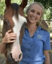 Blonde woman in a blue shirt smiling beside a brown horse with a white blaze on its face.
