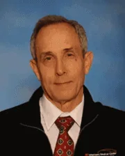 Dr. Jim Schumacher wearing a white shirt, patterned red tie, and dark jacket, against a blue background.