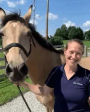 Dr. Naomi Chiero smiling while standing outdoors beside a light brown horse wearing a black halter.
