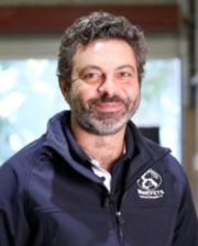 Dr. Nathan Anthony smiling indoors in a navy veterinary jacket with a logo, standing in a softly lit clinical setting.