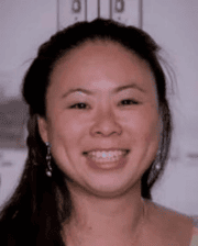 Headshot of a smiling woman with long dark hair, wearing earrings, standing indoors against a blurred background.