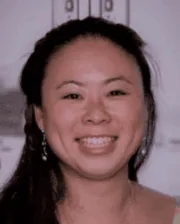 Headshot of a smiling woman with long dark hair, wearing earrings, standing indoors against a blurred background.