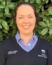 Dr. Rosemary Cuming standing outdoors in front of green foliage, smiling and wearing a navy-blue sweater with a checked shirt.