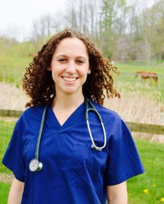 Dr. Sarah Colmer wearing navy scrubs and a stethoscope, smiling outdoors with a grassy field and trees in the background.