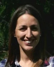 Portrait of a smiling woman with straight brown hair standing outdoors in front of greenery.
