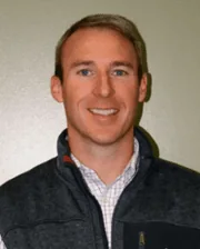 Professional headshot of Dr. Brad Tanner wearing a dark vest over a checkered shirt, smiling against a plain background.