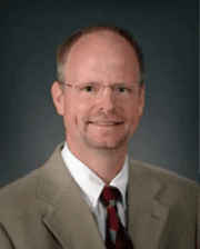 Professional headshot of Dr. Cleet Griffin wearing a tan suit jacket, white shirt, and patterned tie against a dark background.