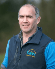 Professional outdoor portrait of Dr. Henry Tremaine wearing a B&W Equine Vets vest and blue shirt, smiling at the camera.