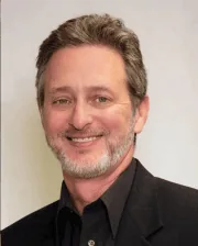 Portrait of Dr. Peter Weinstein smiling, wearing a black blazer and black shirt, with short gray hair and a neatly trimmed beard.