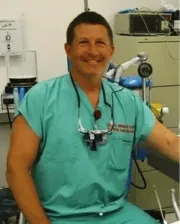 Dr. Stephen Galloway smiling in surgical scrubs, wearing magnifying loupes around his neck, seated in a veterinary surgical room.