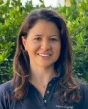Smiling woman with long brown hair standing in front of green foliage, wearing a dark polo shirt.