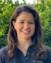 Smiling woman with long brown hair standing in front of green foliage, wearing a dark polo shirt.