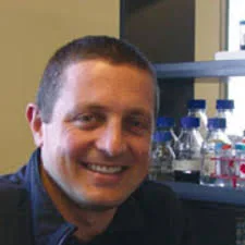 Smiling man with short brown hair seated in a lab setting with scientific bottles on a shelf behind him.
