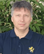 Man with short gray hair wearing a navy polo shirt with a yellow lion emblem, standing in front of green foliage.