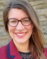 Portrait of a smiling woman with long brown hair, wearing glasses and a red blazer in front of a stone wall.
