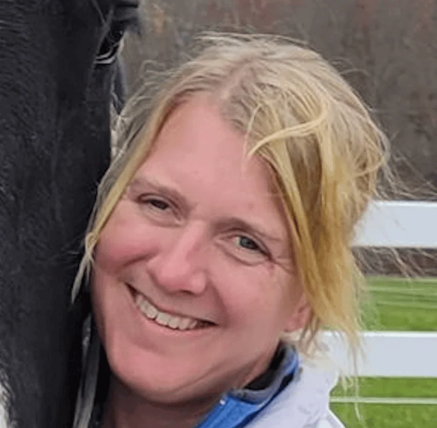 Karin Olsen smiling outdoors beside a black horse with a white fence and autumn trees in the background.