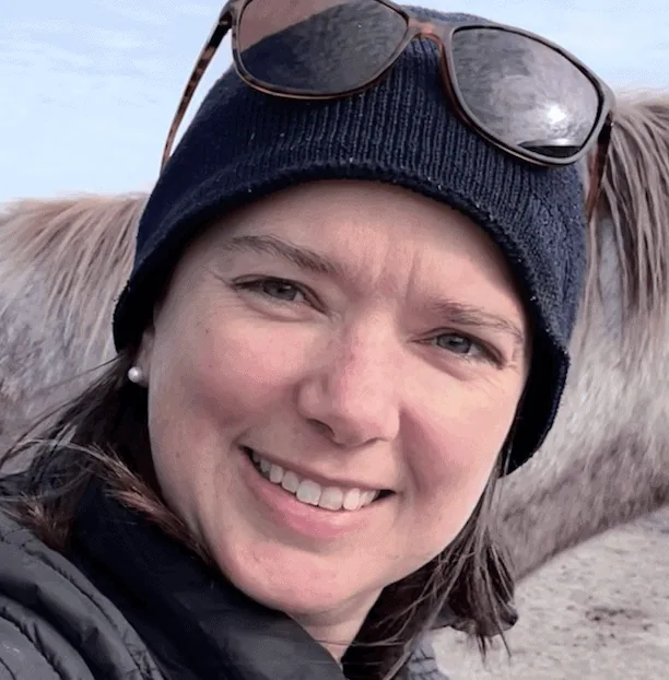 Robyn Thompson smiling outdoors in winter gear with sunglasses on her beanie and a horse in the background.