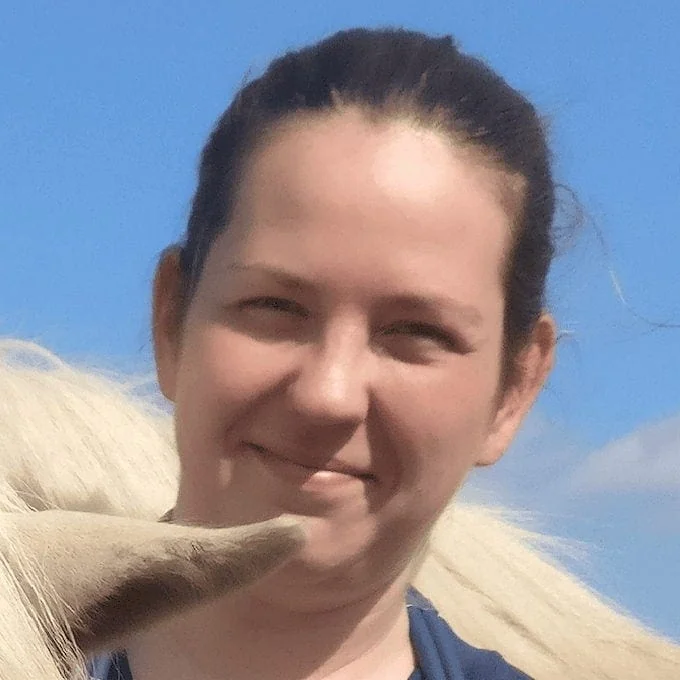 Dr. Sini Maria Peltola smiling outdoors beside a light-colored horse under a clear blue sky.