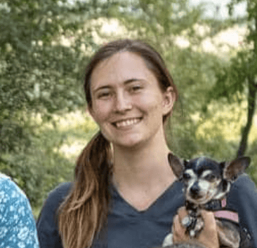 Jandy Torland smiling outdoors in a dark V-neck shirt, holding a small black and tan dog with upright ears.