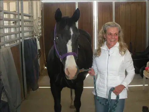 Terry Borreti smiling while holding the lead rope of a black horse with a white blaze, standing together inside a stable.